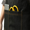 A close-up of a black denim tool apron pocket holding a silver smartphone and two yellow metal rings.