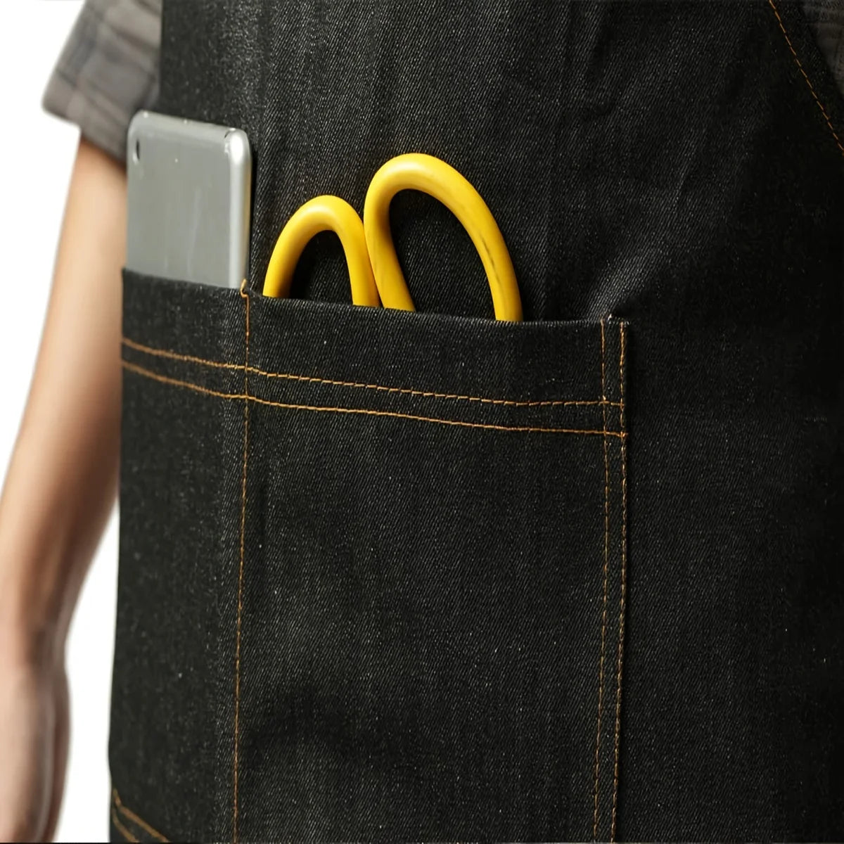 A close-up of a black denim tool apron pocket holding a silver smartphone and two yellow metal rings.