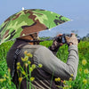 A person wearing a camouflage umbrella hat is taking a photo outdoors in a grassy field.
