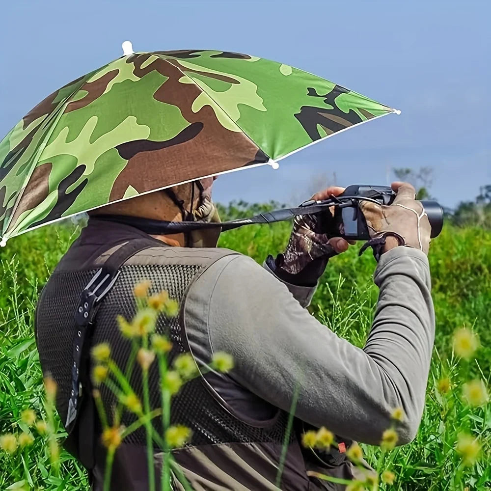 A person wearing a camouflage umbrella hat is taking a photo outdoors in a grassy field.