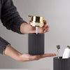 A hand is pouring soap into a black cylindrical dispenser with a gold pump and a toothbrush holder.