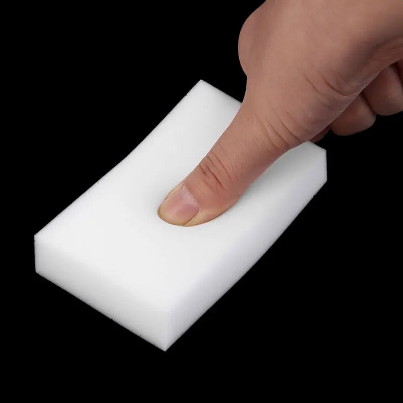 A hand is pressing down on a white melamine cleaning sponge, demonstrating its use as a magic eraser.