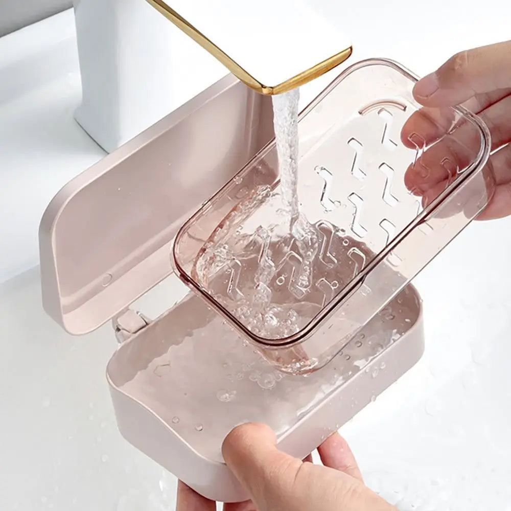 A hand is pouring water into a transparent drainage soap dish, which is part of a pink dispenser.