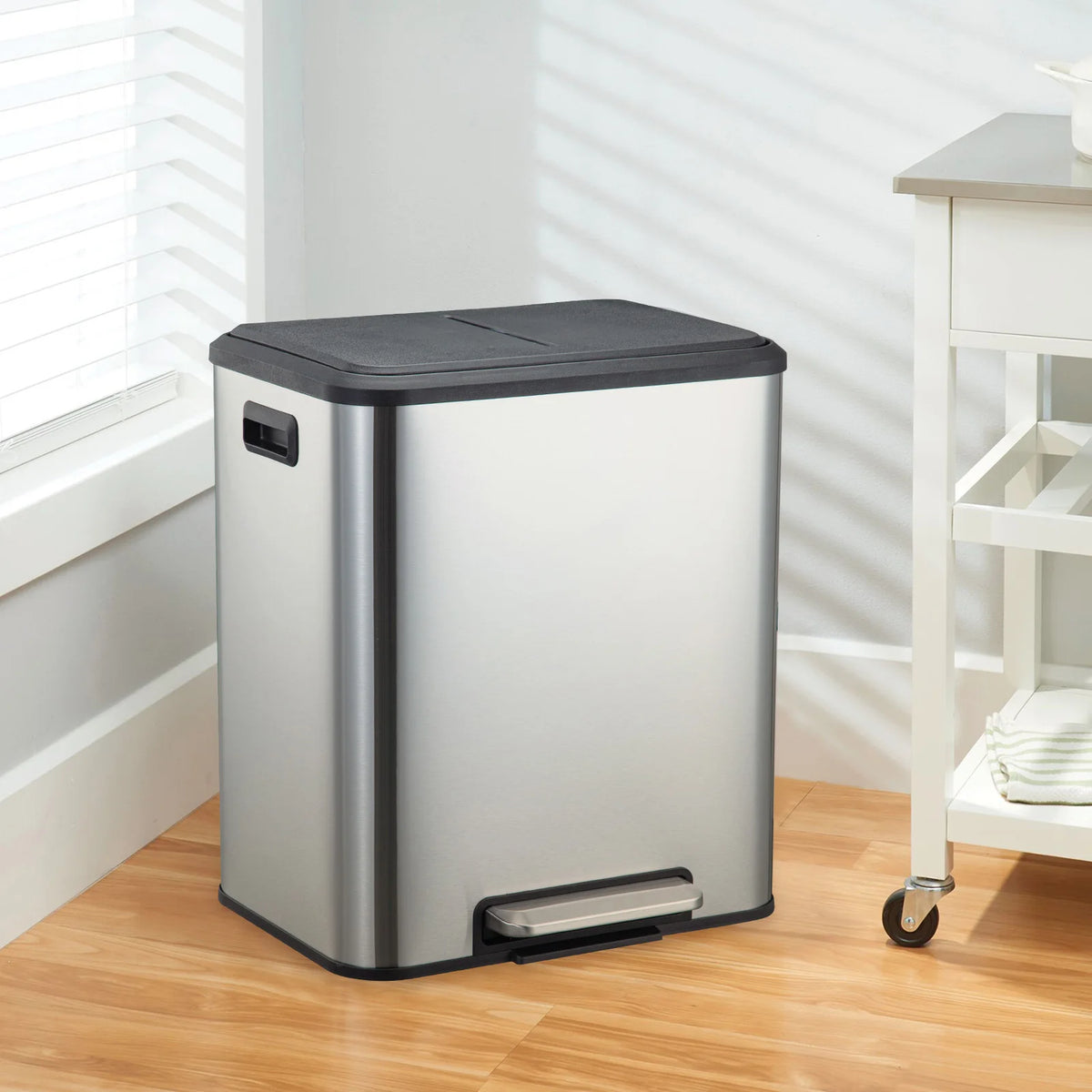 A modern stainless steel dual compartment trash can sits on a wooden floor next to a window.