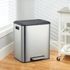 A modern stainless steel dual compartment trash can sits on a wooden floor next to a window.
