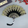 A black and gold folding hand fan with intricate patterns, displayed on a gray surface.
