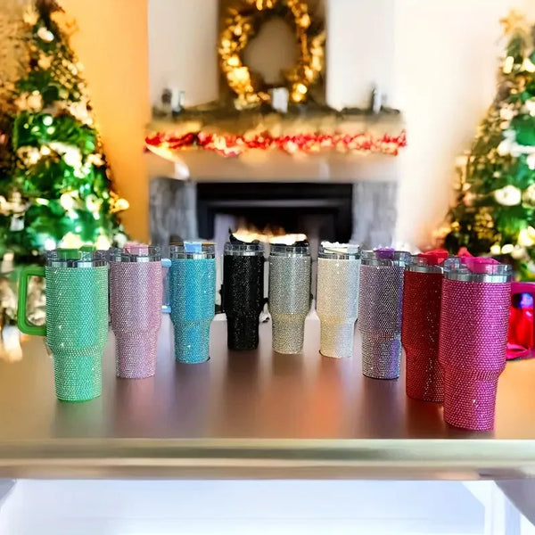 A row of colorful rhinestone stainless steel tumblers displayed on a table in front of a fireplace decorated with greenery.