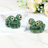 A pair of glossy green ceramic toilet bolt covers resembling small frogs are displayed on a white surface with soft floral gr