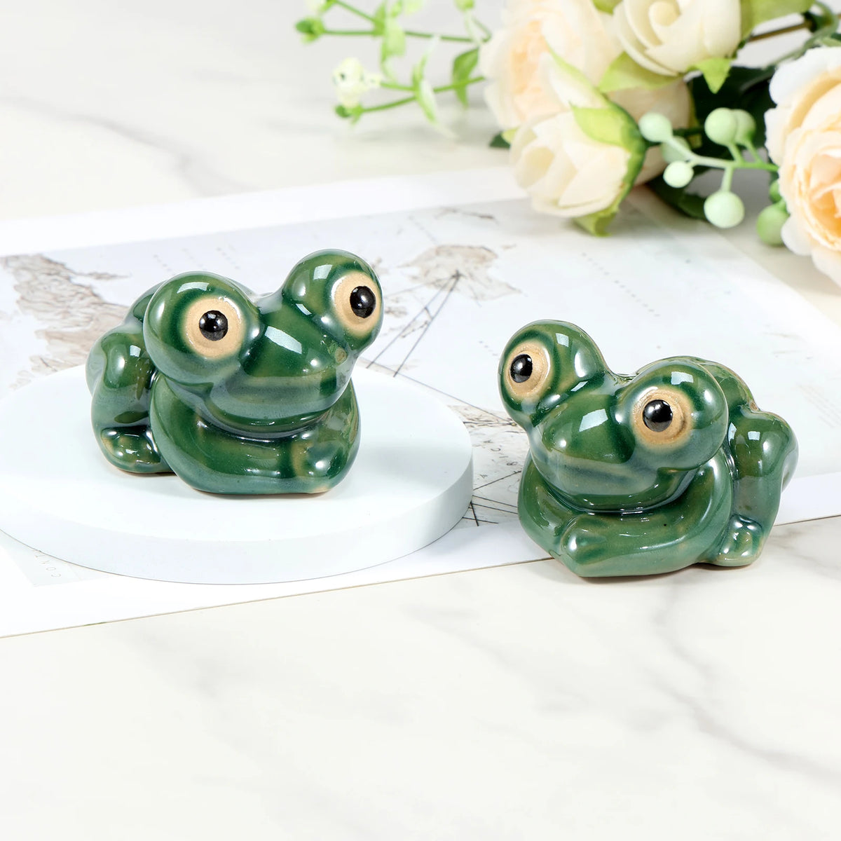 A pair of glossy green ceramic toilet bolt covers resembling small frogs are displayed on a white surface with soft floral gr