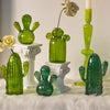Une collection de vases en cactus en verre vert et une bougie blanche sur une surface blanche douce.