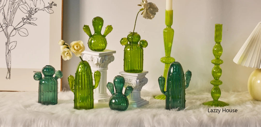 Une collection de vases en cactus en verre vert et une bougie blanche sur une surface blanche douce.