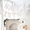 A white banner with pink bows spelling out Happy Birthday hanging against a white wall.