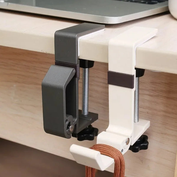 A close-up of an adjustable desk hook bag hanger with a gray clamp and white plastic arms mounted on a wooden surface.