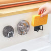 A hand is holding a yellow sponge over a stainless steel holder rack with a suction cup mounted on a white surface.