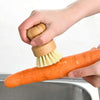 A hand is using a bamboo dish scrub brush with sisal bristles to clean an orange carrot in a metal bowl.