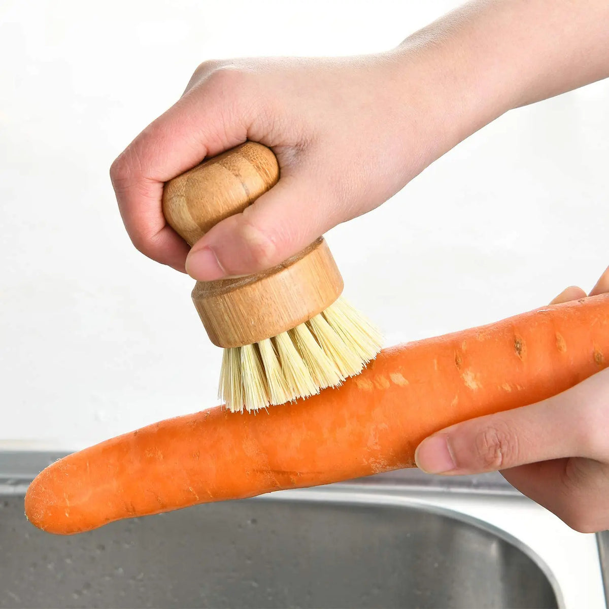 A hand is using a bamboo dish scrub brush with sisal bristles to clean an orange carrot in a metal bowl.