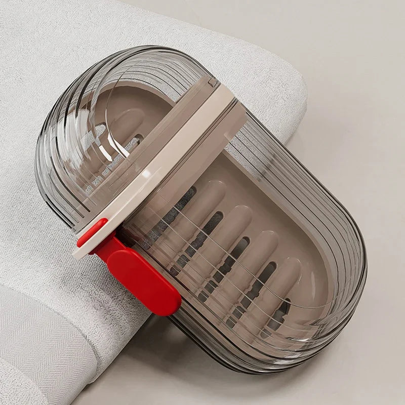Boîte de savon de voyage en verre transparent avec couvercle beige et base rouge, posée sur un tissu blanc.