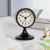 A black round metal alarm clock with a white face and Roman numerals is displayed on a white surface.