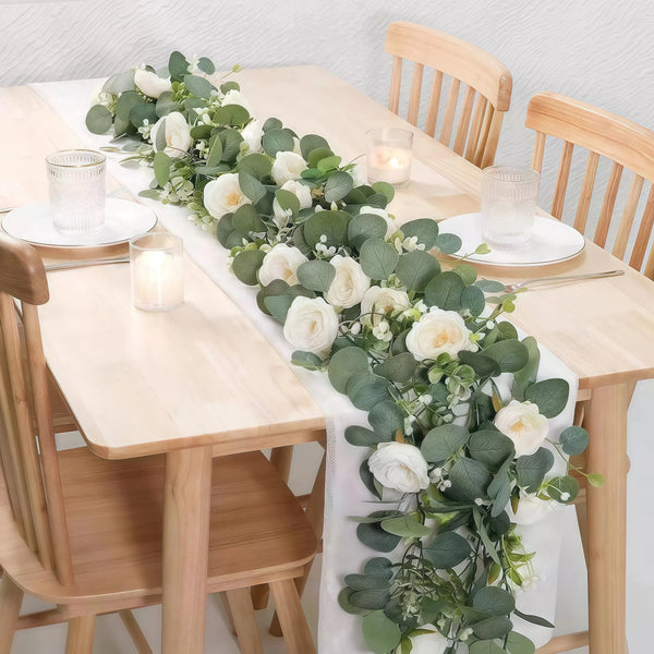 A wooden table decorated with an artificial eucalyptus and rose garland featuring white flowers and green leaves.
