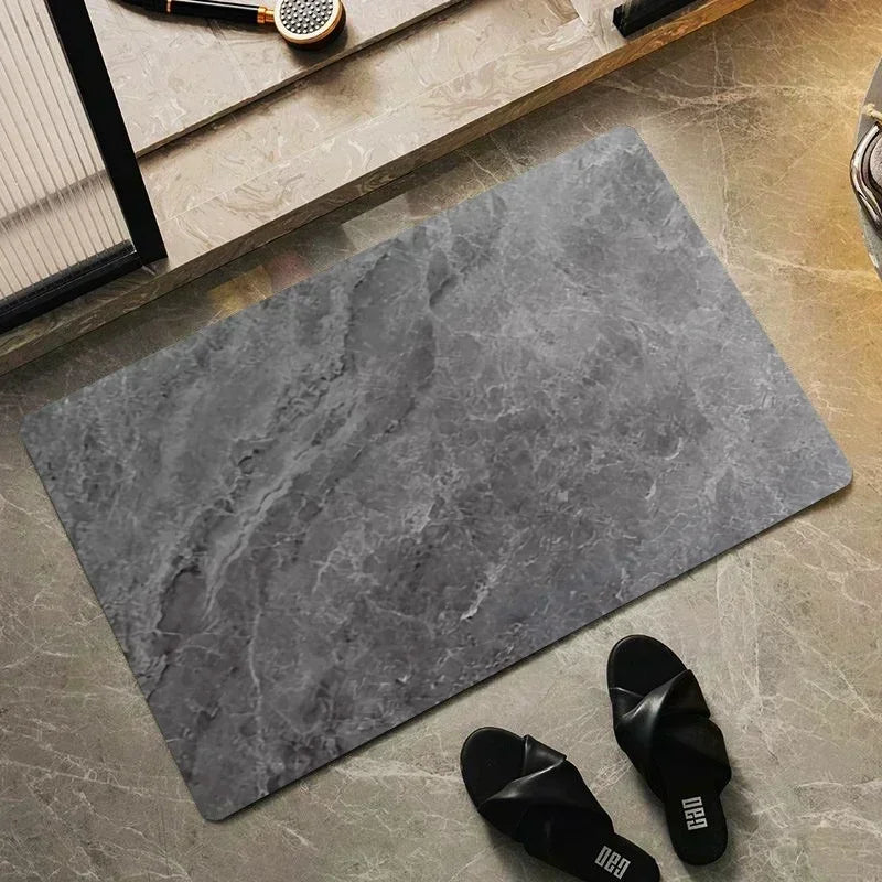 A square gray diatomite bath mat placed on a light stone surface with a black slipper nearby.