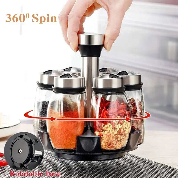 A hand is rotating a set of glass spice jars with metal dispensers on a black rotating base.