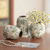 A white ceramic bud vase with green floral patterns sits next to a small silver pearl necklace.