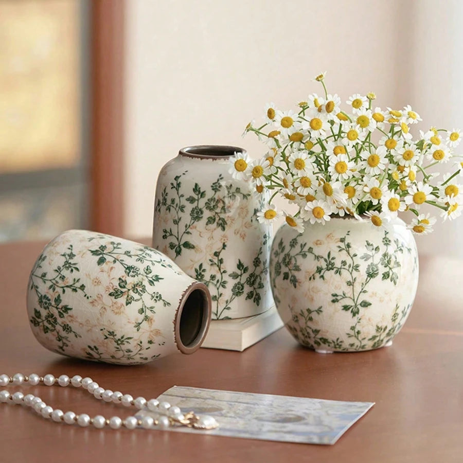 A white ceramic bud vase with green floral patterns sits next to a small silver pearl necklace.