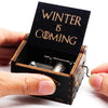 An antique wooden hand crank music box with a dark lid engraved with the text Winter is coming is being held by a hand.