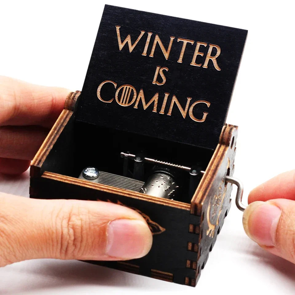 An antique wooden hand crank music box with a dark lid engraved with the text Winter is coming is being held by a hand.