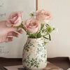 A hand holds a pink ceramic bud vase filled with light pink roses and green foliage.