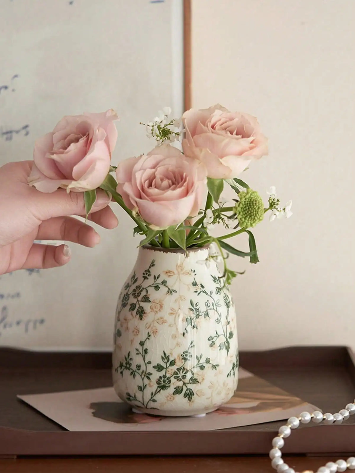 A hand holds a pink ceramic bud vase filled with light pink roses and green foliage.