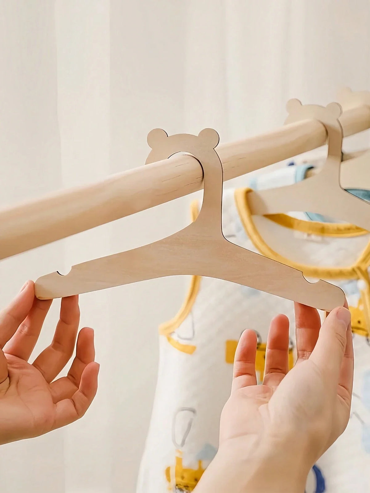 A pair of wooden clothing hangers for kids are being held up against a white curtain.