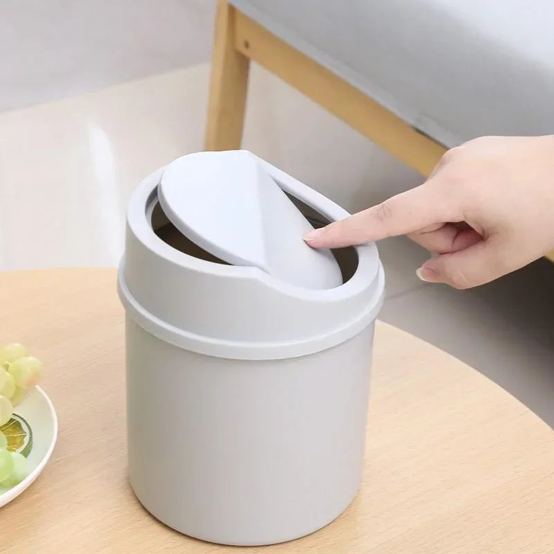 A hand is flipping the lid of a compact, light gray desktop trash can with a flip cover.
