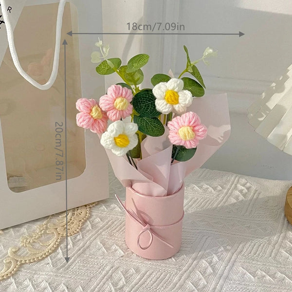 A bouquet of pink and white artificial yarn flowers displayed in a pink paper cup on a white textured surface.