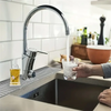 Eine Hand hält ein Glas Wasser, das durch einen modernen Küchenhahn in eine Edelstahlspüle fließt.