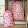 A pink drawstring storage bag with a red cherry pattern is displayed in a wooden cabinet.