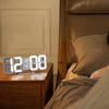 A white digital alarm clock displaying 12:00 is resting on a wooden nightstand next to a brown leather headboard.