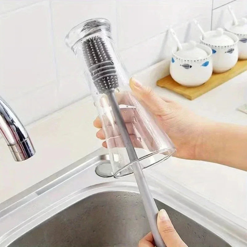 A hand holds a silicone bottle brush with a long handle and bristles over a kitchen sink.
