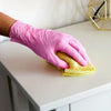A hand wearing a pink disposable nitrile glove is holding a yellow cloth on a white countertop.