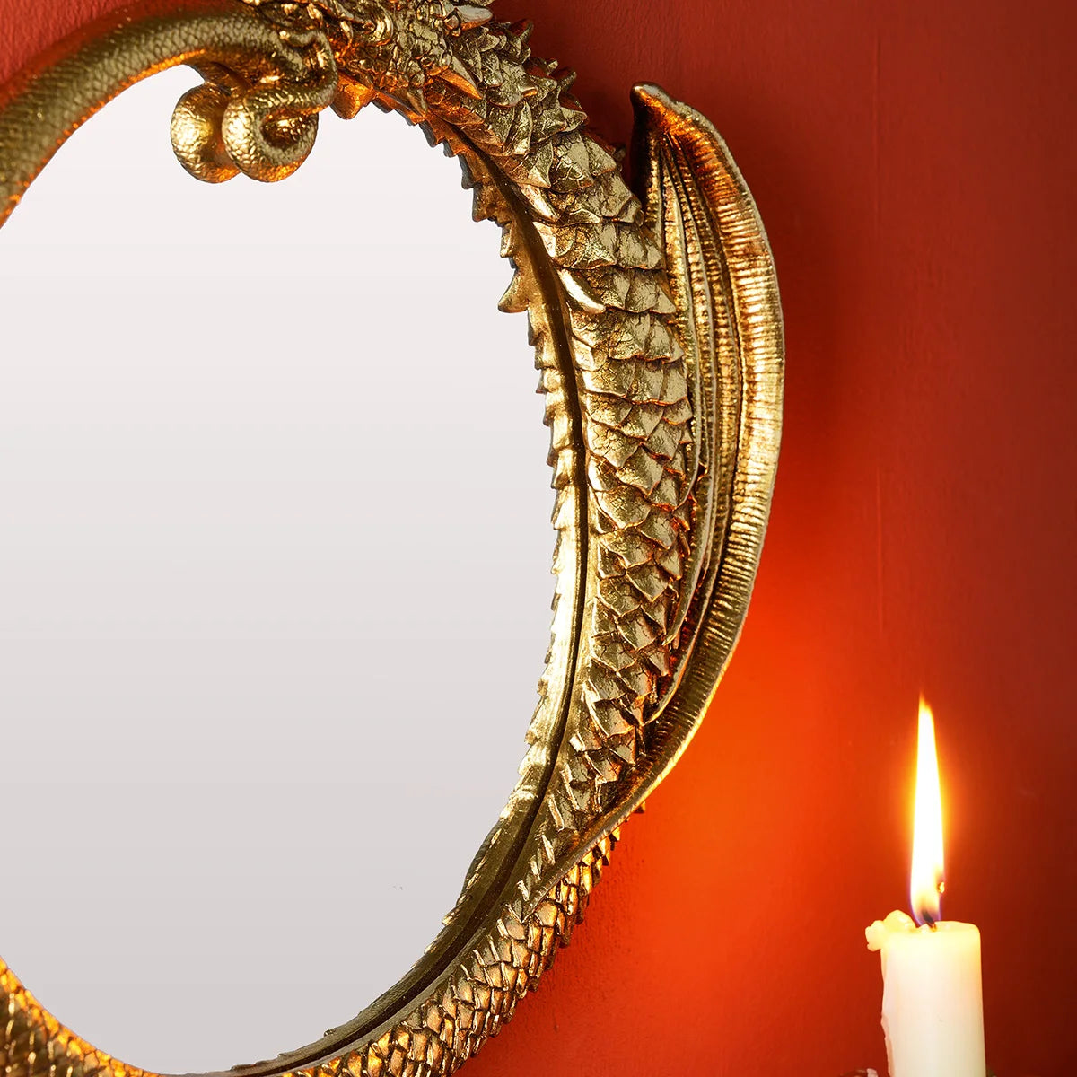 A close-up of a round, ornate gold-framed mirror reflecting a warm orange wall and a lit white candle.