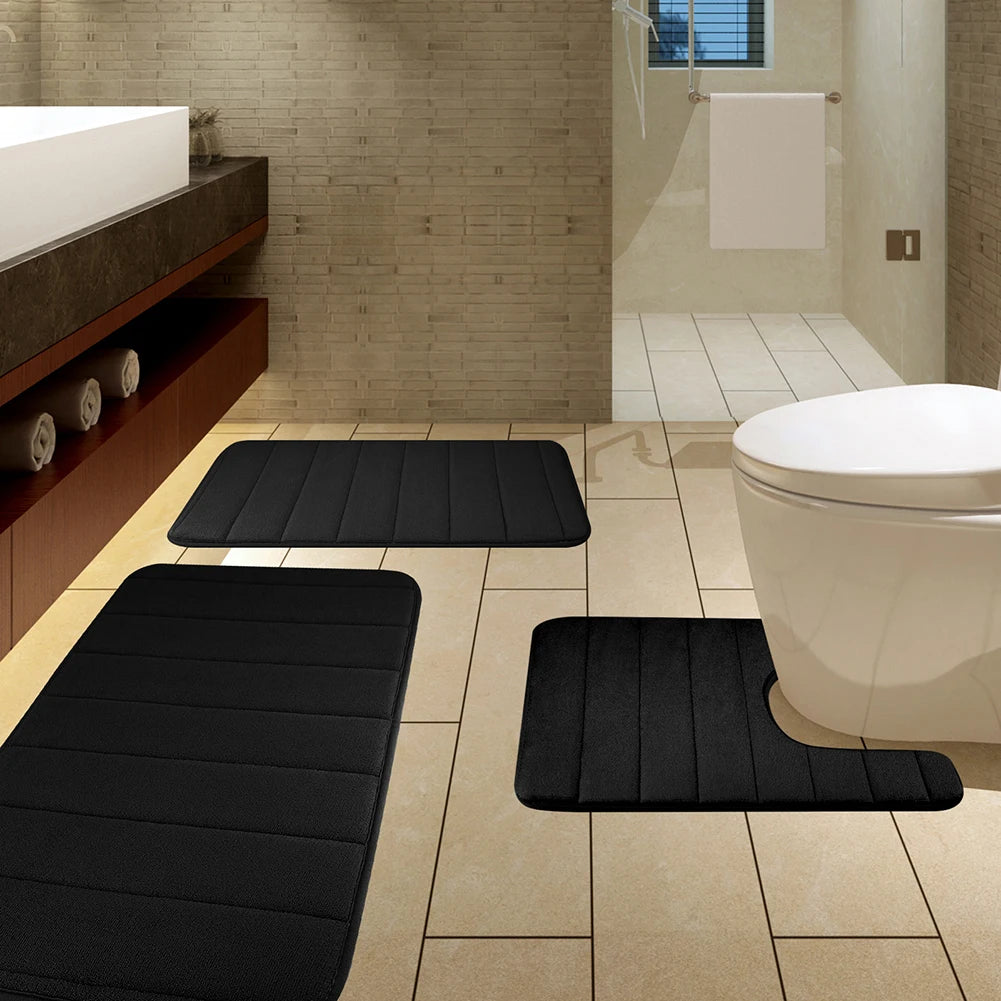 Ein Set aus drei schwarzen Memory-Schaum-Badematten auf einem Fliesenboden neben einer weißen Toilette.
