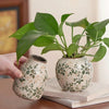 A hand holds a ceramic bud vase with green leaves, featuring a floral pattern, resting on a stack of books.