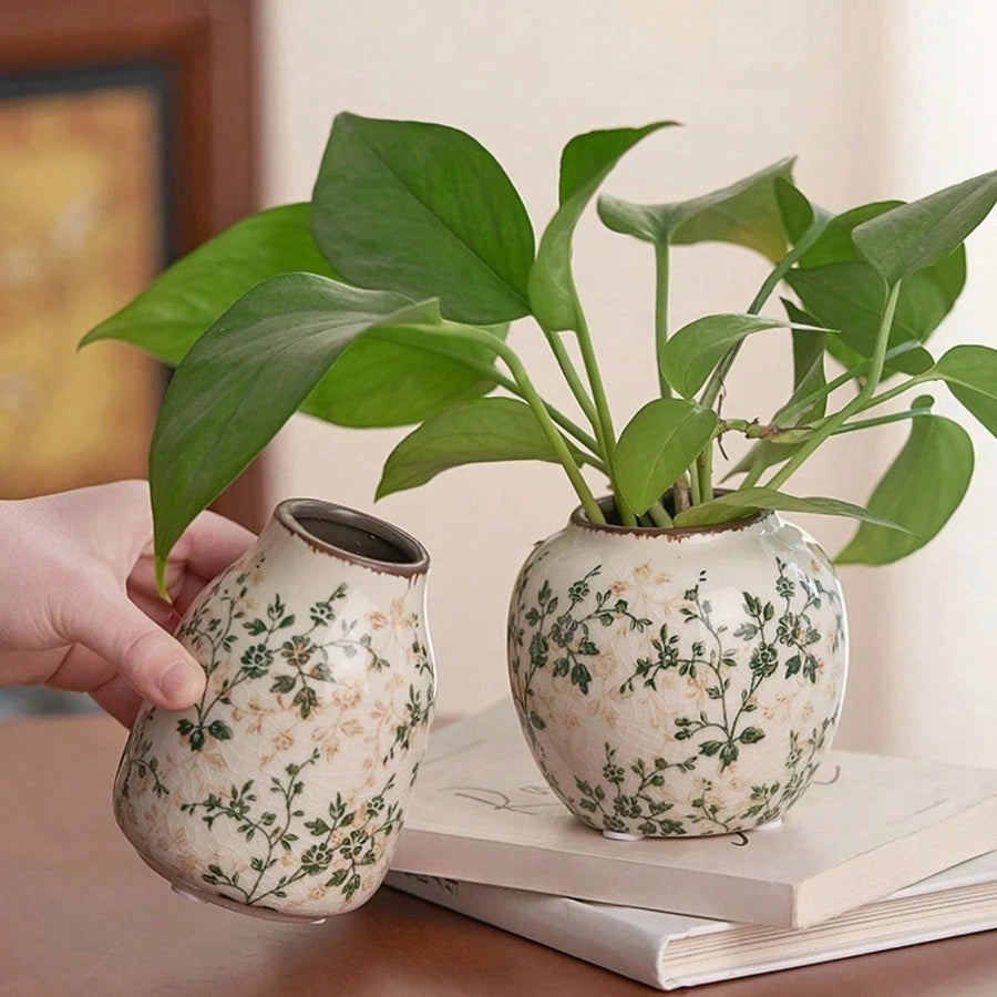 A hand holds a ceramic bud vase with green leaves, featuring a floral pattern, resting on a stack of books.