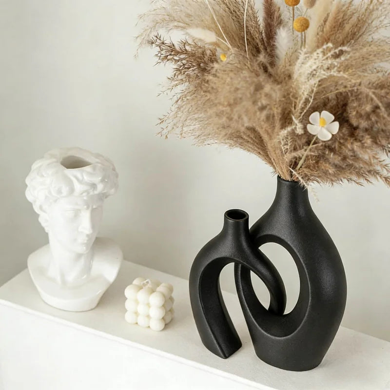 A black ceramic vase holds dried beige floral arrangement and a white plaster bust on a white shelf.