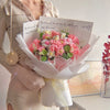 Un bouquet de fleurs de carnation en coton fait main, emballé dans du papier blanc et tenu par une personne.