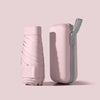 A light pink compact 5-fold travel umbrella with a gray strap is shown against a pale background.