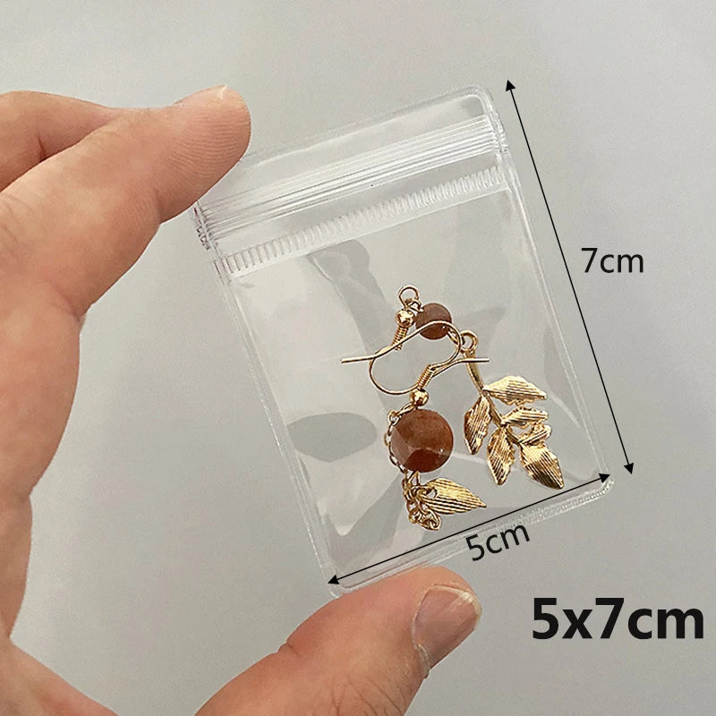 A hand holds a clear PVC zip lock jewelry pouch containing a pair of gold-toned leaf and bead earrings.