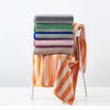 A stack of plaid cotton bath towels in various muted colors, including gray, green, blue, and pink, next to an orange striped