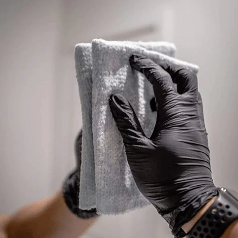 A hand wearing a black nitrile cleaning glove is holding and wiping a gray cloth or towel.