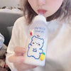 A person holds a white water bottle featuring a blue outline drawing of a bear and a small yellow fruit.